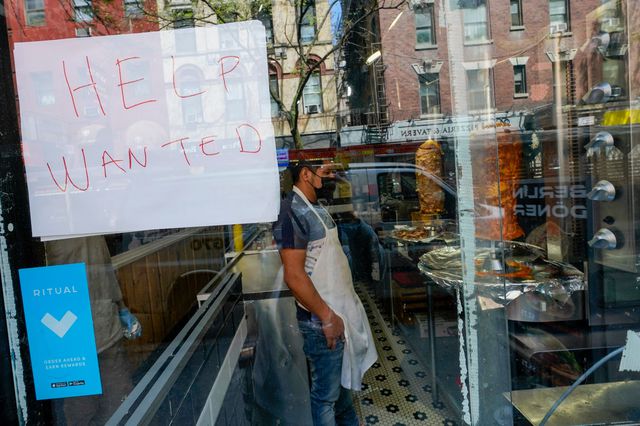A sign saying "Help wanted" in the window of a restaurant, with worker seen through the glass
